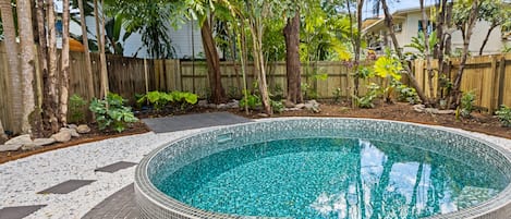 A circular pool with a tiled edge, surrounded by a wooden deck and a variety of tropical plants.