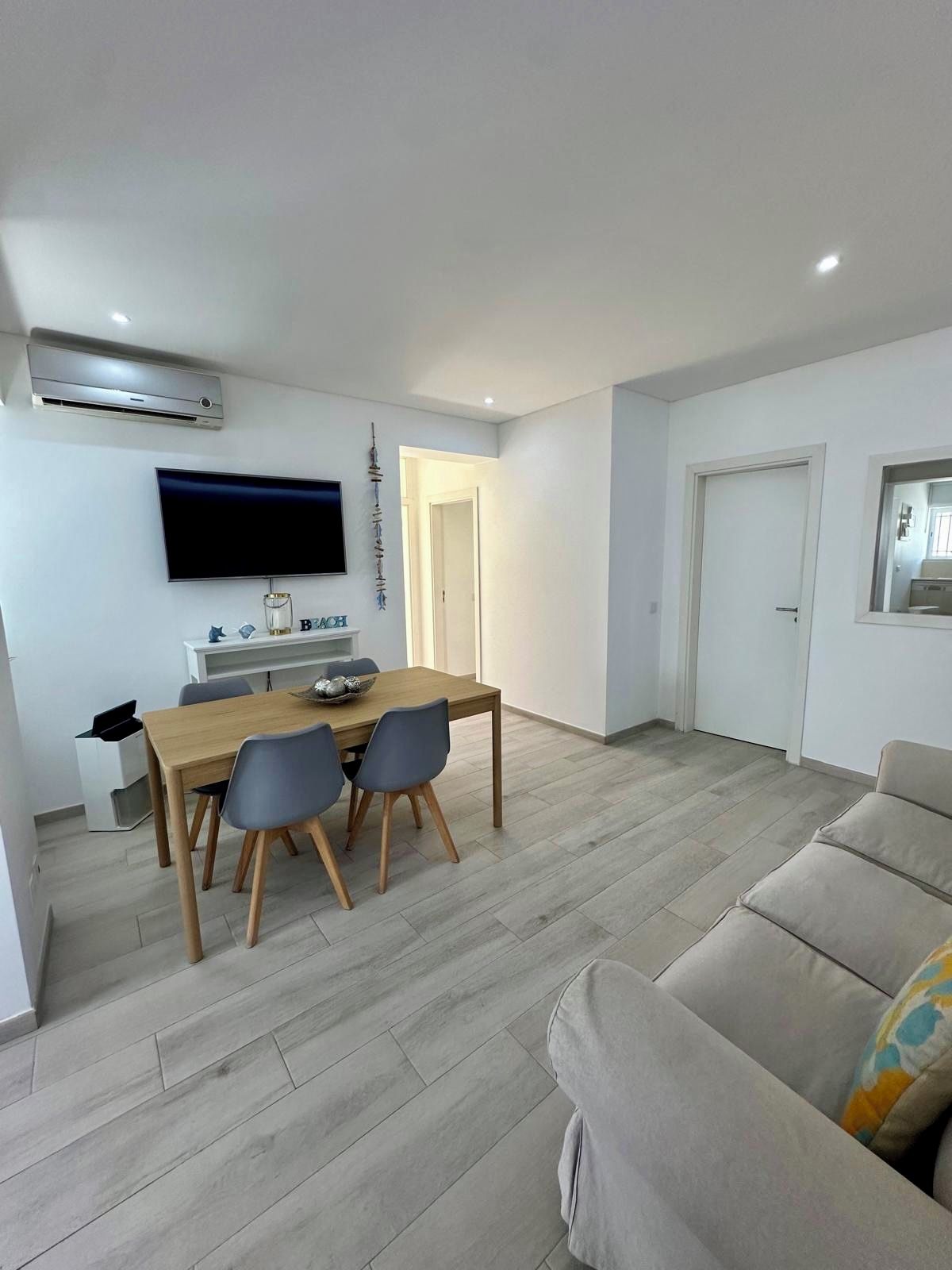 A modern living room with a dining area, a flat-screen TV mounted on the wall, and a sofa.