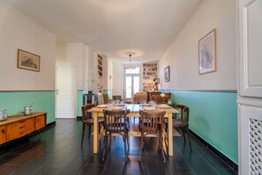 Ein Essbereich mit einem langen Tisch, gedeckt für sechs Personen, umgeben von Stühlen. Links befindet sich ein Bücherregal voller Bücher, und ein Durchgang zu einem weiteren Raum.