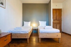 Ein Zimmer mit zwei Holzbetten, jeweils mit weißer Bettwäsche und Kissen, einem Nachttisch mit Lampe und einer Holztür mit Kreuz.