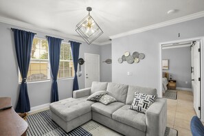 Ein modernes Wohnzimmer mit einem grauen Ecksofa, einer geometrischen Pendelleuchte und einem gemusterten Teppich.