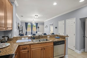 A modern kitchen with granite countertops, wooden cabinets, a dishwasher, and a window with curtains. - Waterstone Paradise | Your 5 star Florida Stay (Davenport)