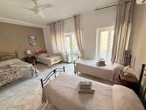 Quarto de hotel com duas camas, um ventilador de teto, uma janela com cortinas e uma pequena mesa com abajur.