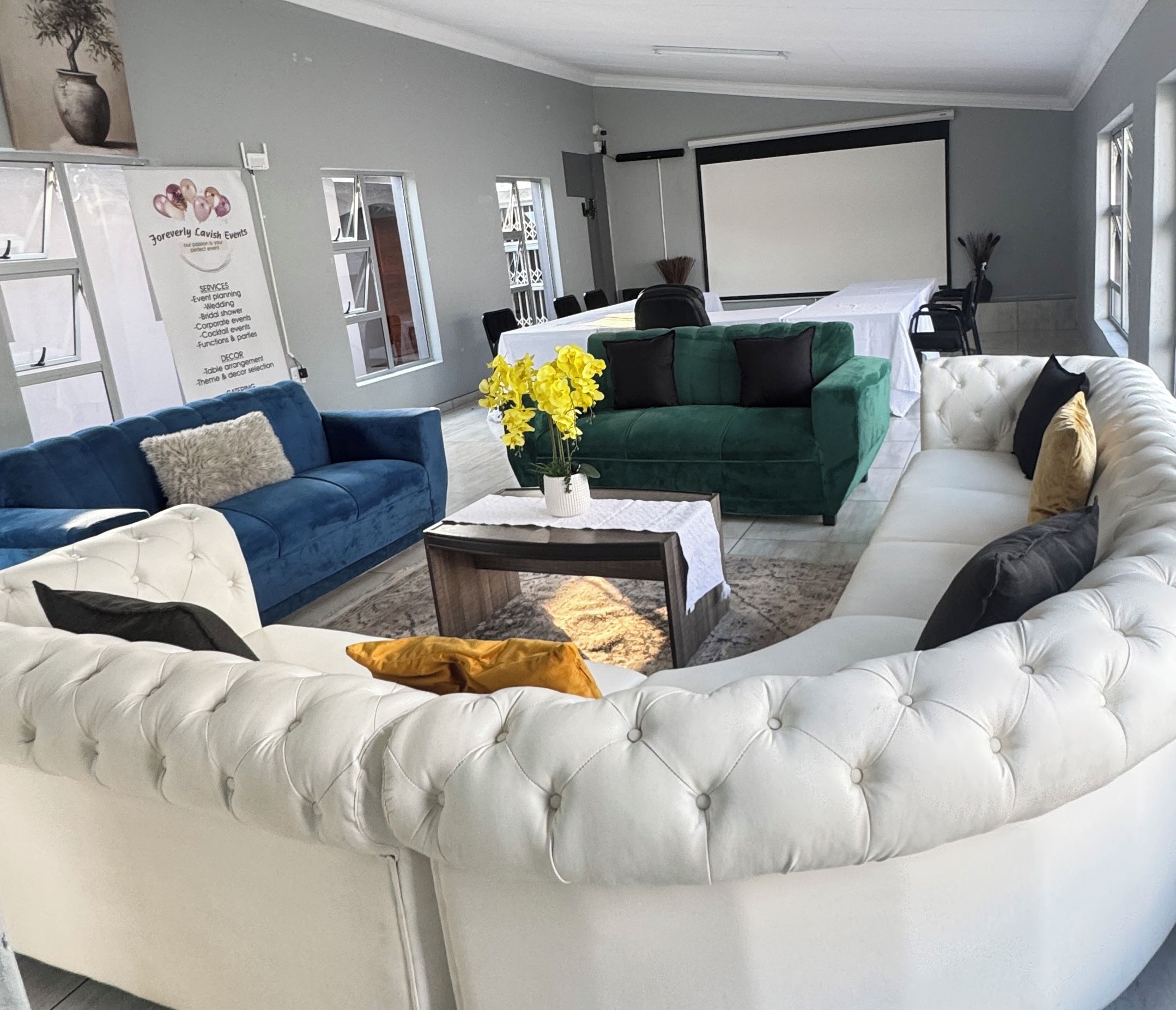 Een moderne woonkamer met een gebogen witte bank, een blauwe bank en een groene bank. Er staat een salontafel met een vaas vol gele bloemen.