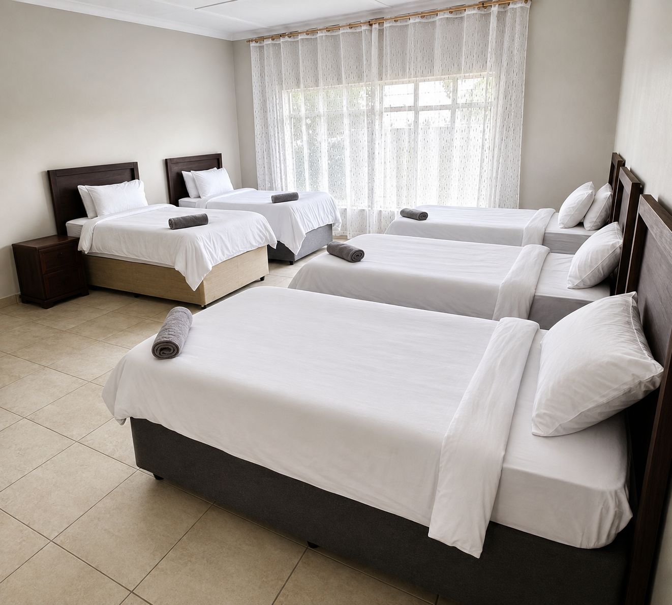 Habitación de hotel con cuatro camas individuales, cabecera de madera, mesita de noche y un amplio ventanal con cortinas traslúcidas.