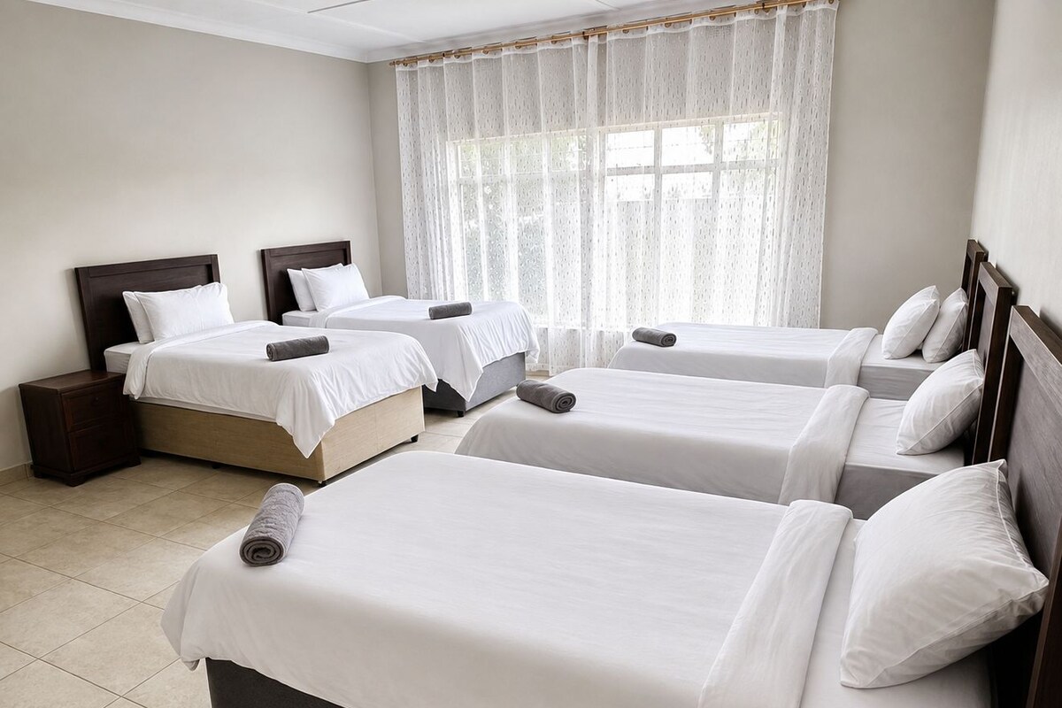 A hotel room with four single beds, a wooden headboard, a nightstand, and a large window with sheer curtains.