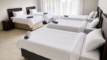 Habitación de hotel con cuatro camas individuales, cabecera de madera, mesita de noche y un amplio ventanal con cortinas traslúcidas.