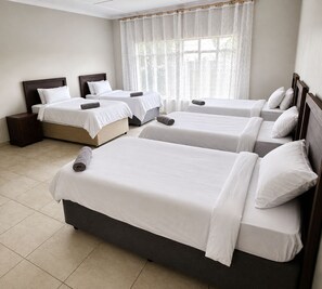 Habitación de hotel con cuatro camas individuales, cabecera de madera, mesita de noche y un amplio ventanal con cortinas traslúcidas.