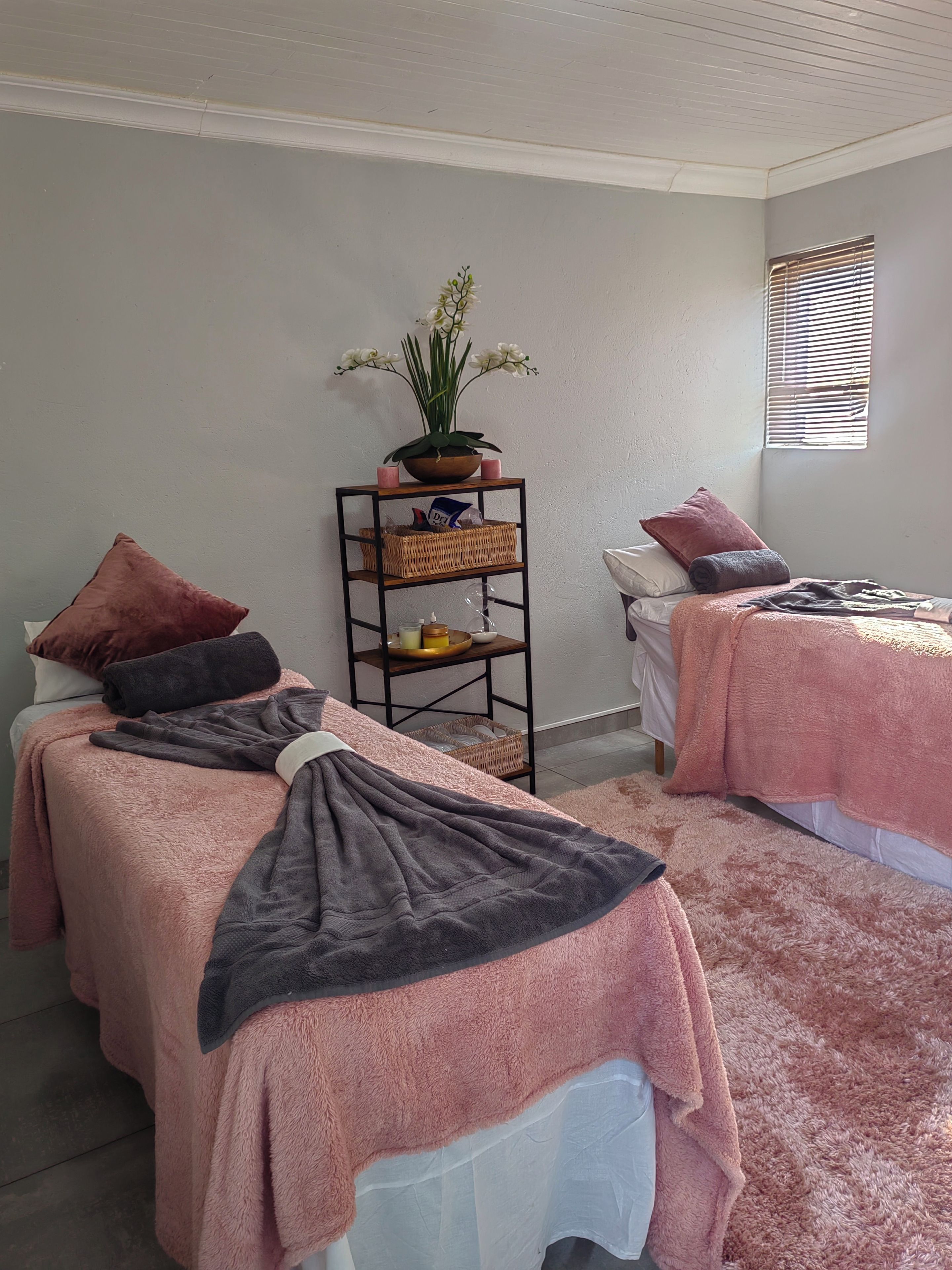 Een kamer met twee bedden, elk met een roze handdoek, een klein plankje met een plantje en een raam met jaloezieën.