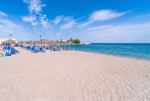 Une plage bordée de rangées de transats et de parasols bleus, un ciel dégagé et une mer calme.