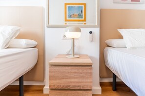 Une chambre d’hôtel avec deux lits, une table de chevet avec une lampe, une petite table de nuit en bois, un thermostat fixé au mur et un tableau encadré au-dessus du miroir.