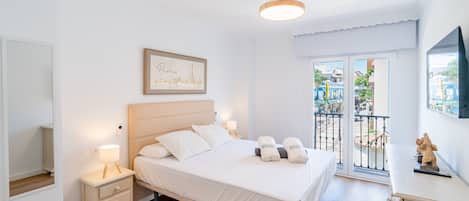 Una habitación de hotel con cama, mesitas de noche, un cuadro enmarco de París, un balcón con vista a la calle y un televisor montado en la pared.