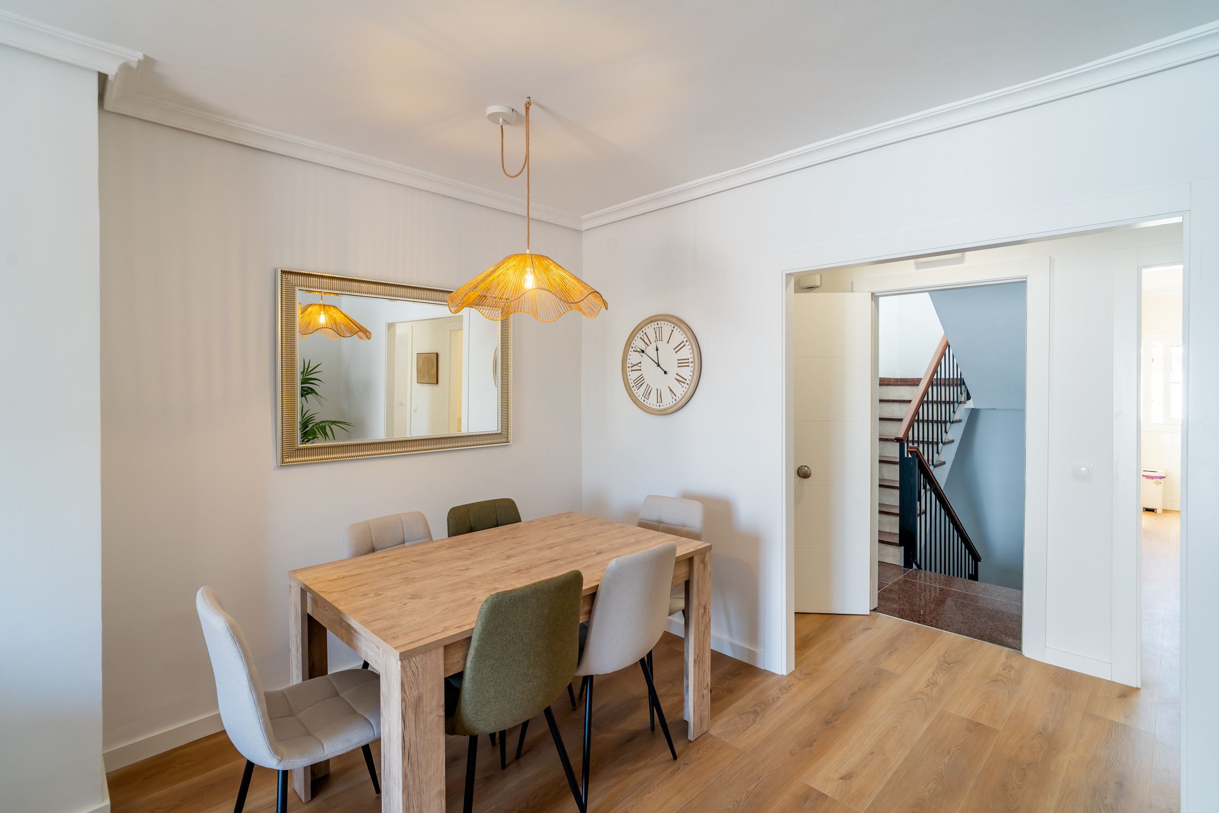 Un coin repas avec une table et des chaises en bois, une horloge murale et un escalier menant à un autre niveau.