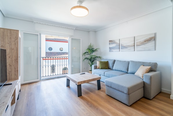 Una sala moderna con un sofá gris, una mesa de centro de madera y un ventanal con vistas a un edificio tradicional.