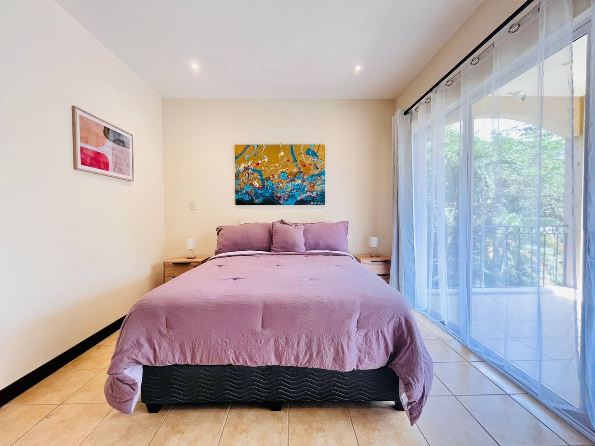 A bedroom with a large bed, a nightstand, a painting, and a view through a sliding door.