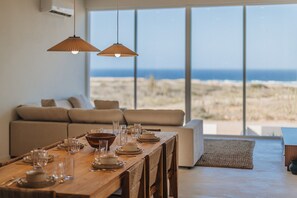 A dining area with a long wooden table set for a meal, a sofa, and a view of the beach.