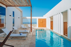 Un área de piscina con sillones, una pérgola de madera y vista al mar.