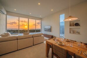A modern interior with a large window overlooking a sunset, a dining area with a wooden table set, and a cozy seating area with a sofa.