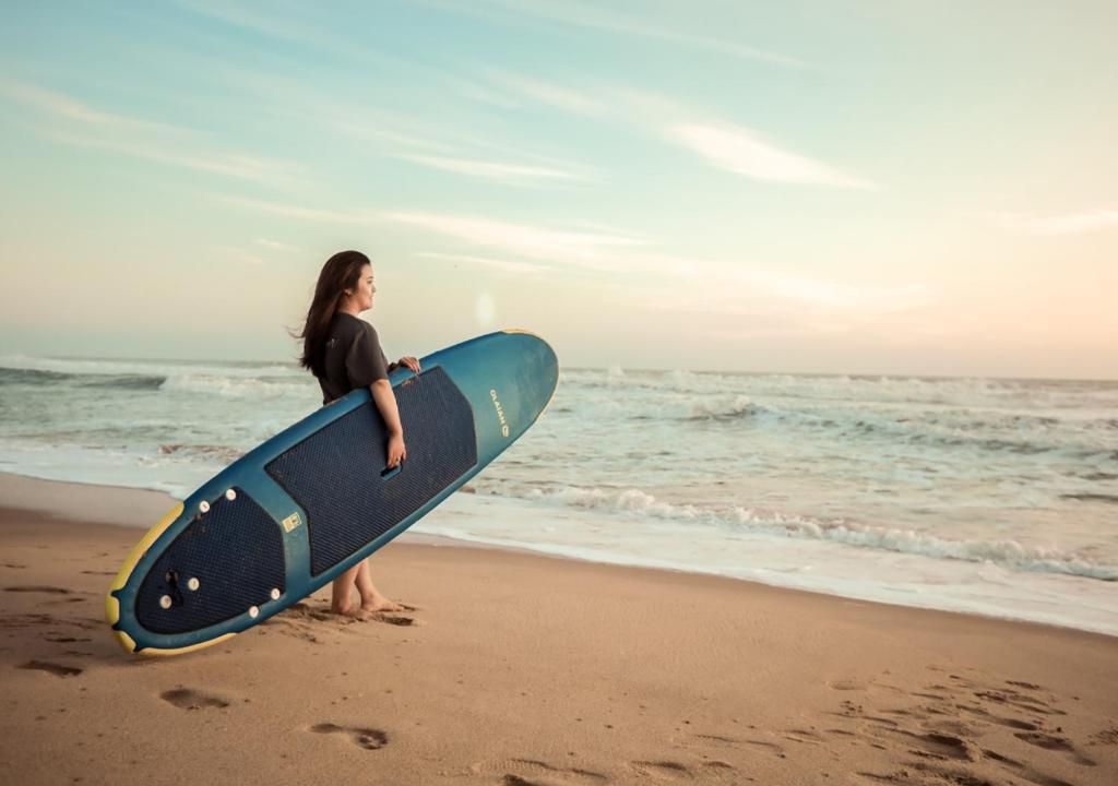 Una persona sostiene una tabla de surf en la playa, con el océano al fondo.