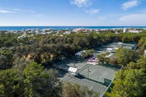 Ein Tennisplatz, umgeben von dichter Vegetation, mit freiem Blick auf das Meer und eine ferne Skyline.