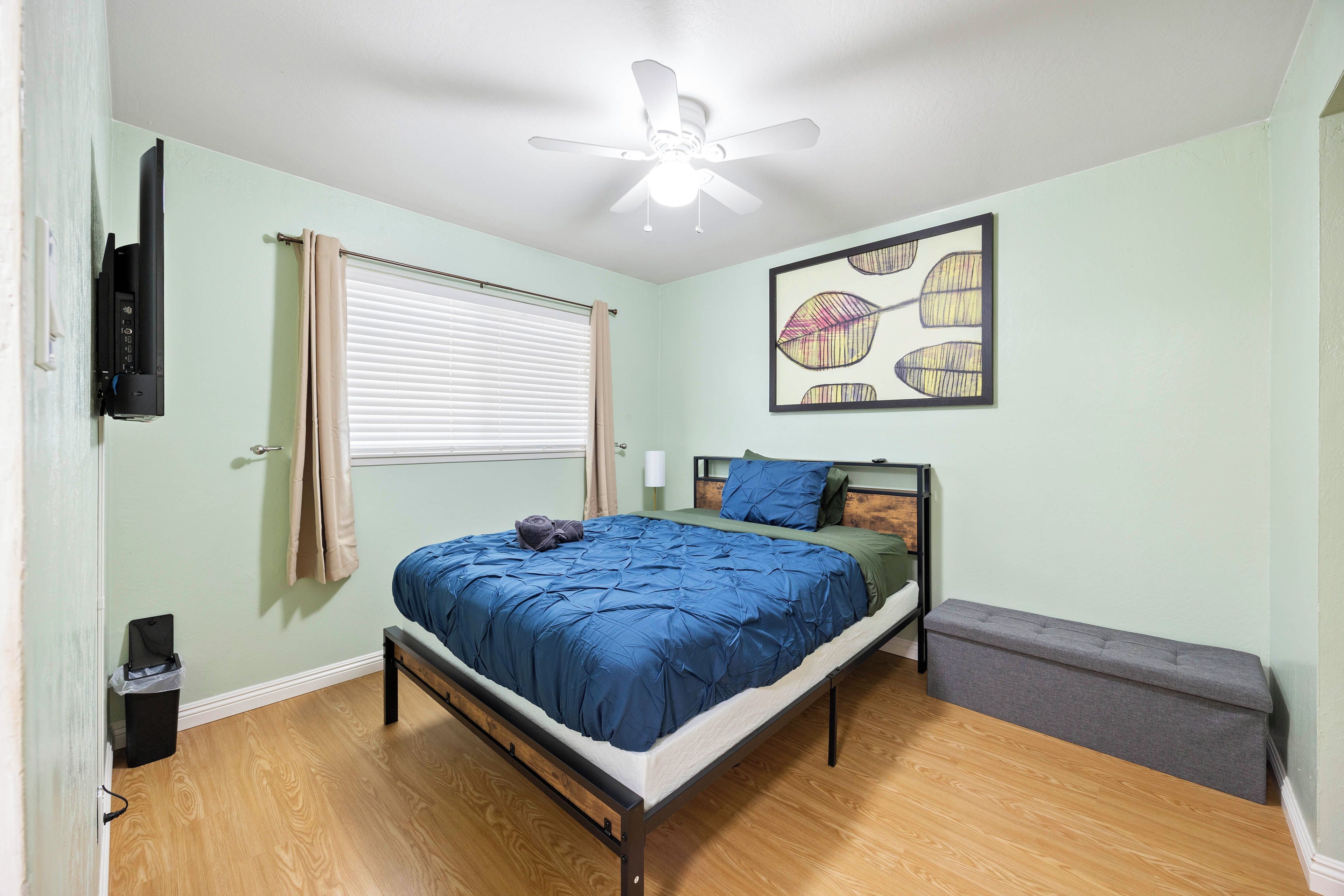 Quarto com cama, ventilador de teto, janela com persianas, quadro decorativo e um banquinho.