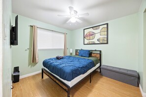 Quarto com cama, ventilador de teto, janela com persianas, quadro decorativo e um banquinho.