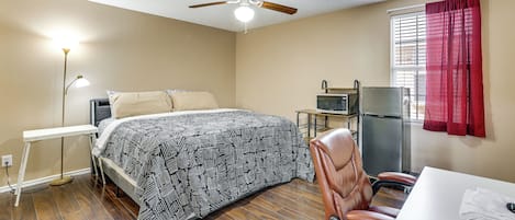 Quarto com cama, escrivaninha, cadeira, ventilador de teto, janela com cortinas e geladeira.