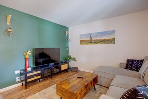 Una sala de estar con un televisor de pantalla plana, una mesa de centro de madera, un sofá gris y un cuadro de un paisaje desértico.