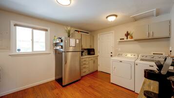 A kitchen with a stainless steel refrigerator, a built-in oven, a washing machine, and a dryer. There is a window with blinds, a door, and a shelf with plants.