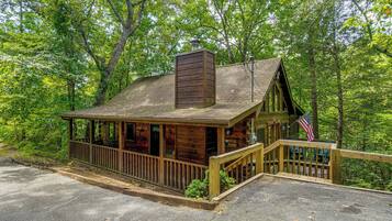A rustic cabin with a porch and a chimney, surrounded by dense forest.