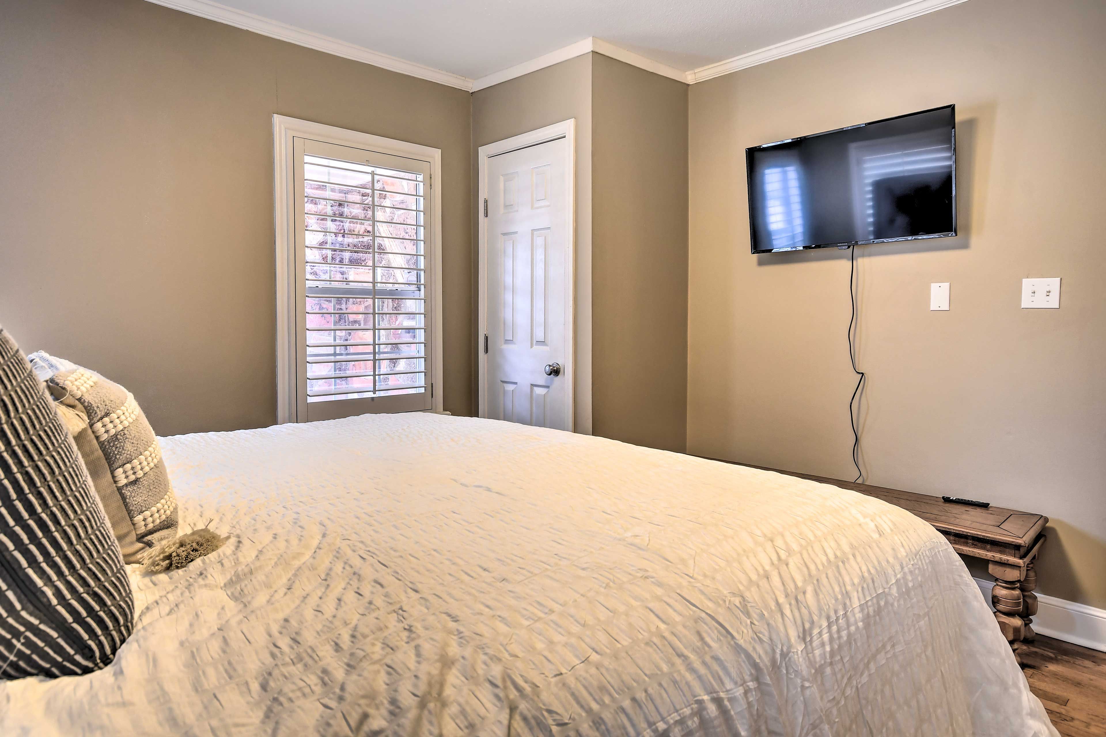 Un dormitorio con una cama grande, televisor montado en la pared y ventana con persianas.