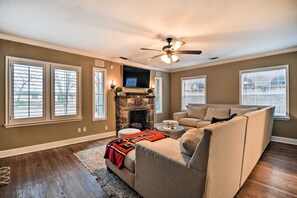 A living room with a stone fireplace, a ceiling fan, and a large window with blinds. - Walk to Texas Tech! Upscale Family Retreat (Lubbock)