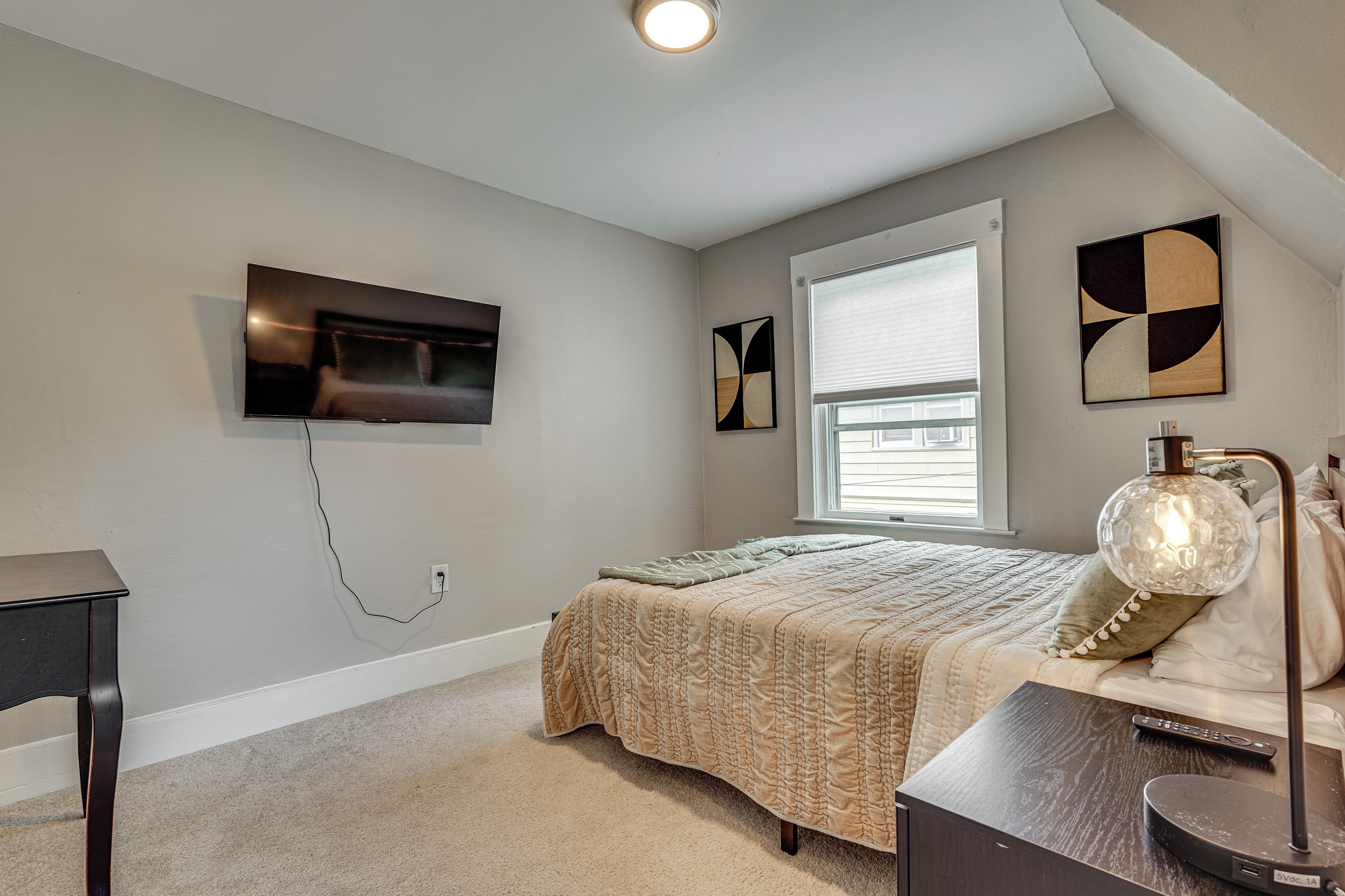 Un dormitorio con una cama, televisión montada en la pared, una ventana y una lámpara.