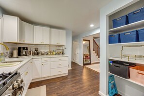 A modern kitchen with white cabinetry, a gas stove, and a coffee maker. There is a wooden floor and a hallway leading to a staircase. - Extended Stay Near Yale: East Haven Home w/ Yard! (New Haven)