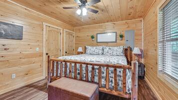 A wooden-paneled room with a bed, a ceiling fan, a bench, and a window with blinds.