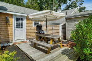 Um deck de madeira com mesa de piquenique e churrasqueira, cercado por um portão de madeira e uma casa com porta e janelas brancas.