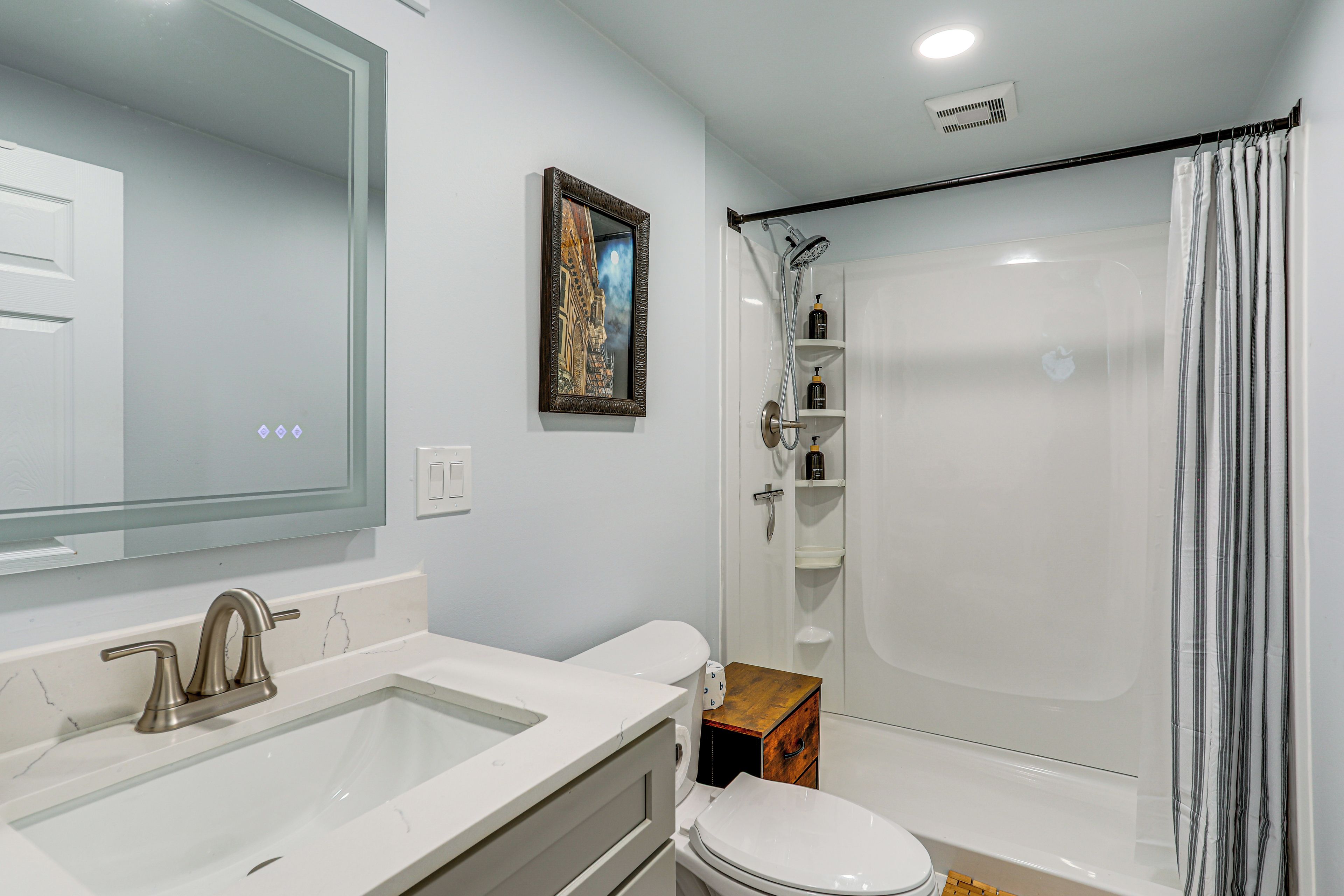 A bathroom with a sink, mirror, toilet, shower, and a shelf with toiletries.