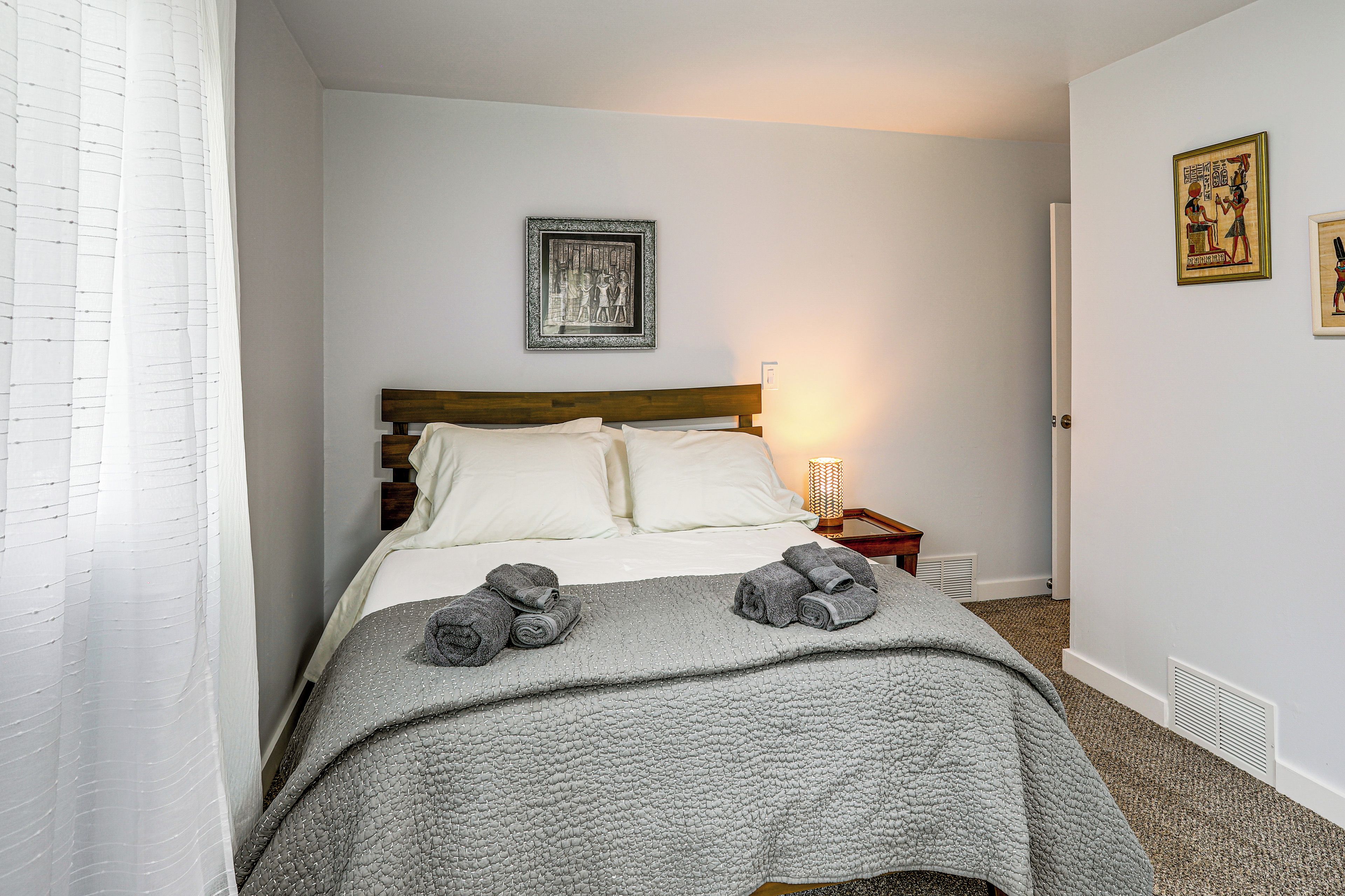 A neatly made bed with a grey quilt and pillows, a bedside lamp, and two framed pictures on the wall.