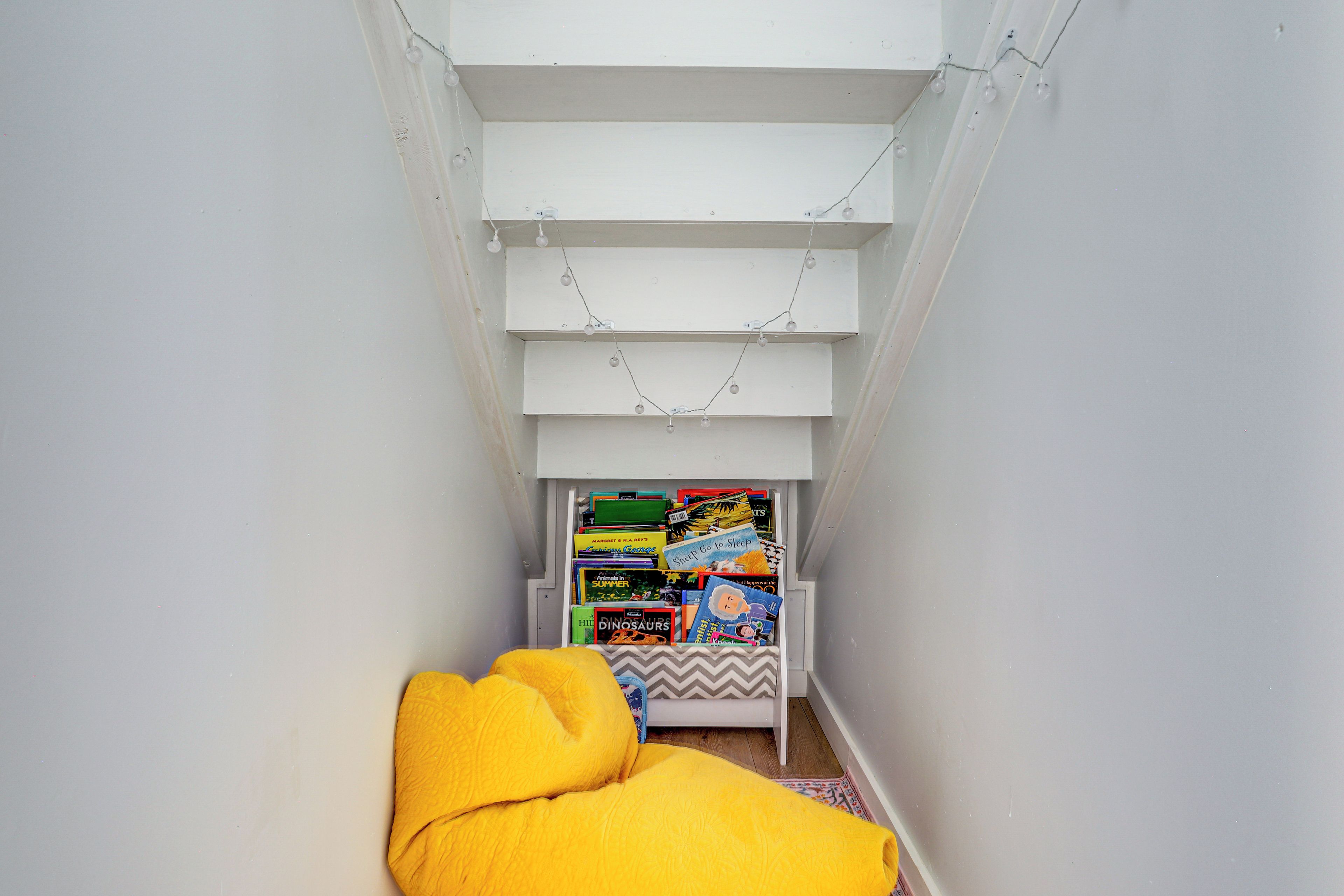 A narrow room with a sloped ceiling, a bed with a yellow blanket, and a bookshelf filled with books.