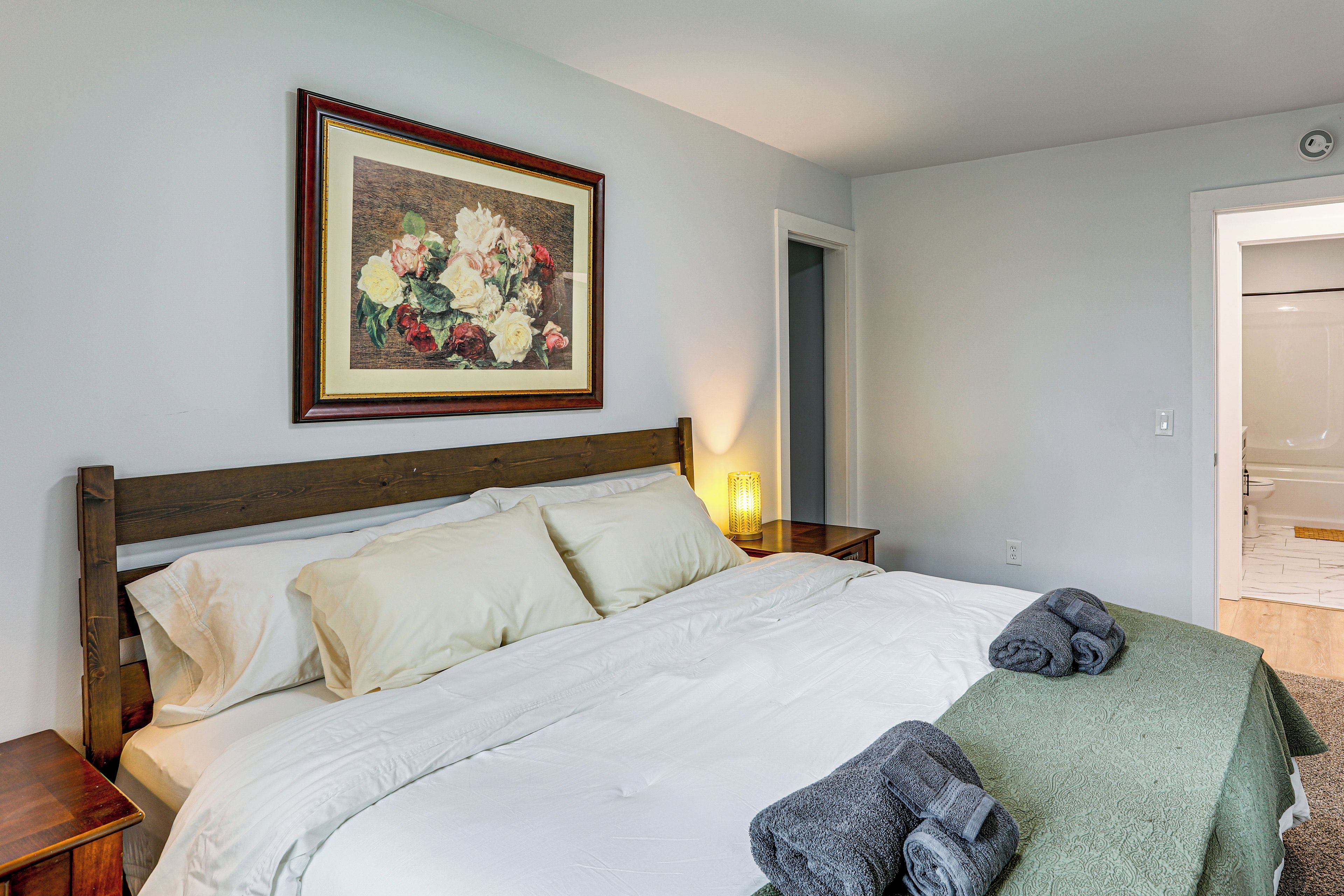 Un dormitorio con una cama de madera, ropa de cama blanca, una obra de arte floral enmarcada, una lámpara de noche y una toalla verde.