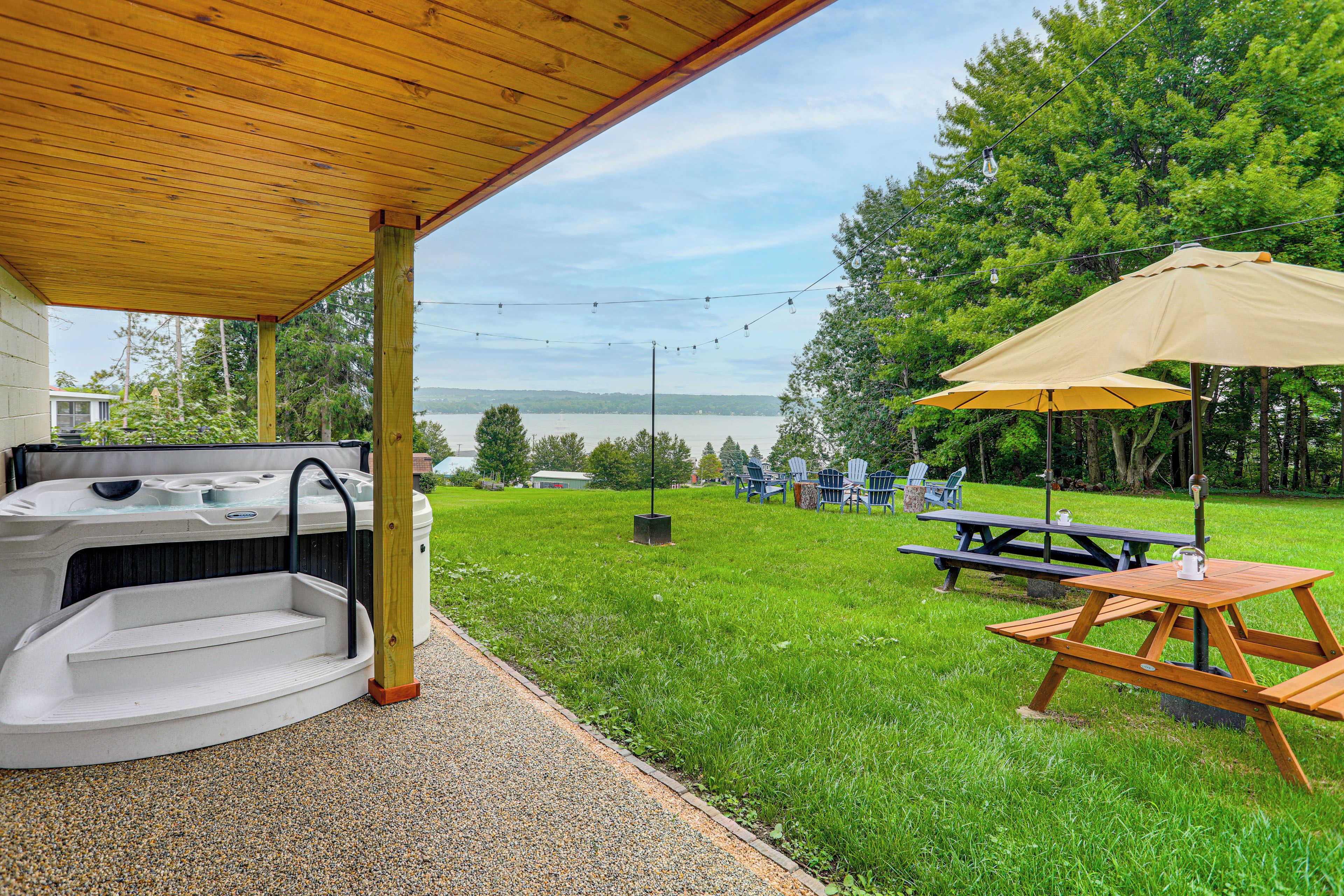 Une terrasse couverte avec un jacuzzi, une table de pique-nique et un parasol, donnant sur une zone herbeuse avec des arbres et un plan d'eau en arrière-plan.
