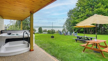 A covered patio with a hot tub, picnic table, and umbrella, overlooking a grassy area with trees and a body of water in the background.