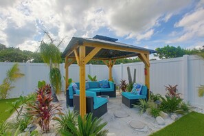 A covered outdoor seating area with two sofas and a table, surrounded by tropical plants and a white fence.