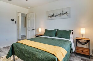 Un dormitorio con una cama, dos mesitas de noche con lámparas, una puerta y un cuadro en la pared que representa un paisaje urbano.
