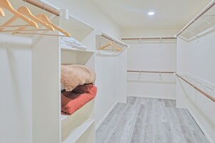 Un closet tipo pasillo con perchas y estantes de madera, una cama con una colcha rosa, y piso de madera.