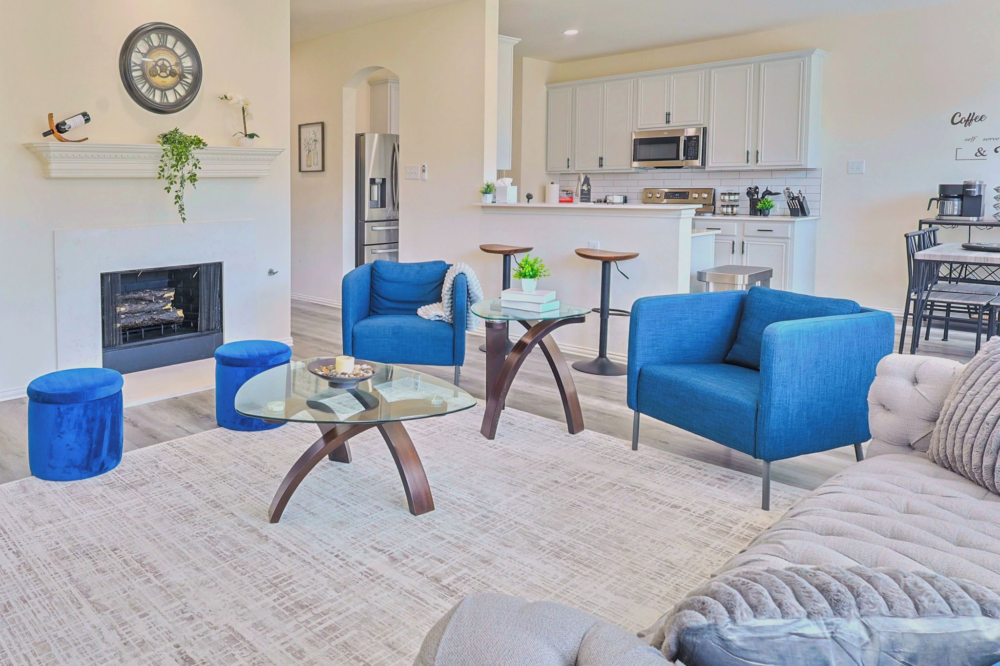 A modern living room with a fireplace, blue armchairs, a glass coffee table, and a kitchen in the background.