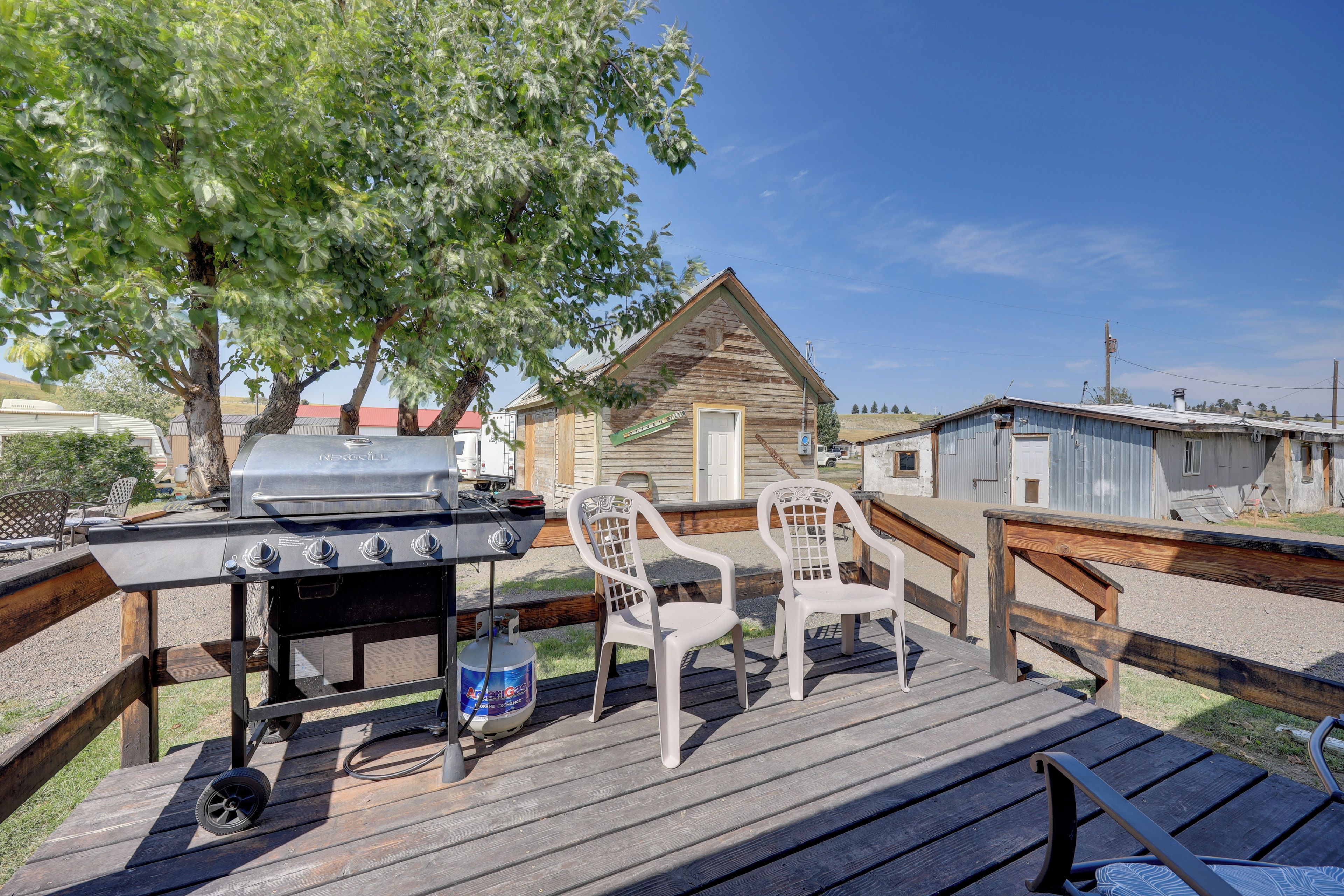 A wooden deck with a barbecue grill and chairs, a small wooden building, and a clear blue sky.