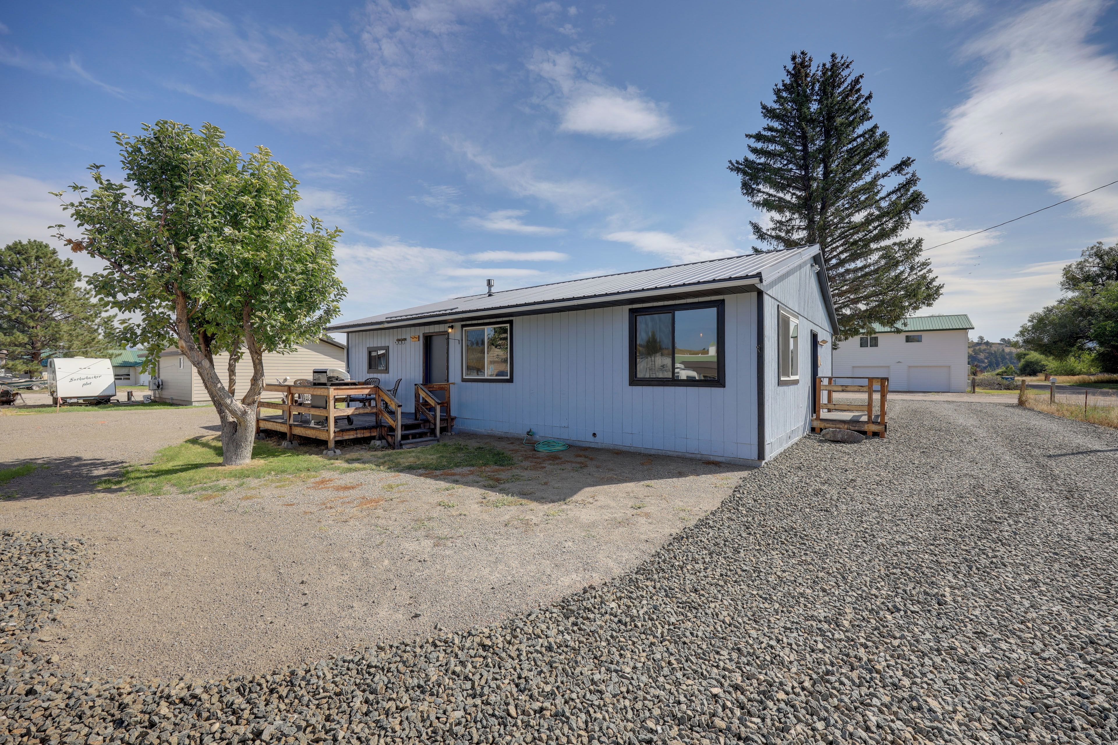 A single-story house with a gravel driveway, a wooden deck, and a large tree in front.