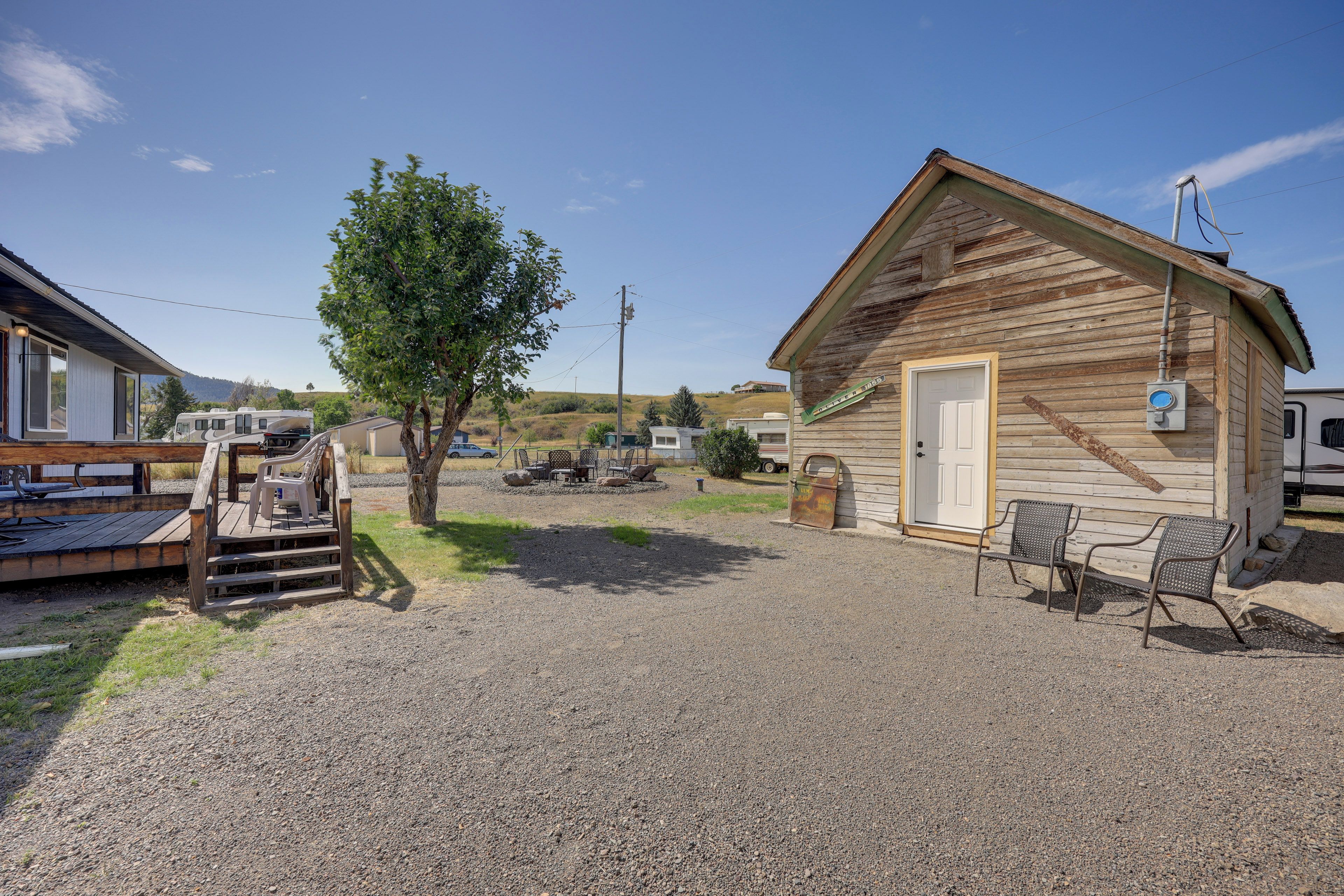 A gravel driveway leading to a wooden cabin with a door and a window, a metal chair, and a small table with chairs outside.