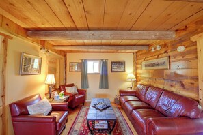 Interior acolhedor de uma cabana, com paredes de madeira, um sofá de couro vermelho, uma poltrona combinando, uma mesa de centro e uma lareira.